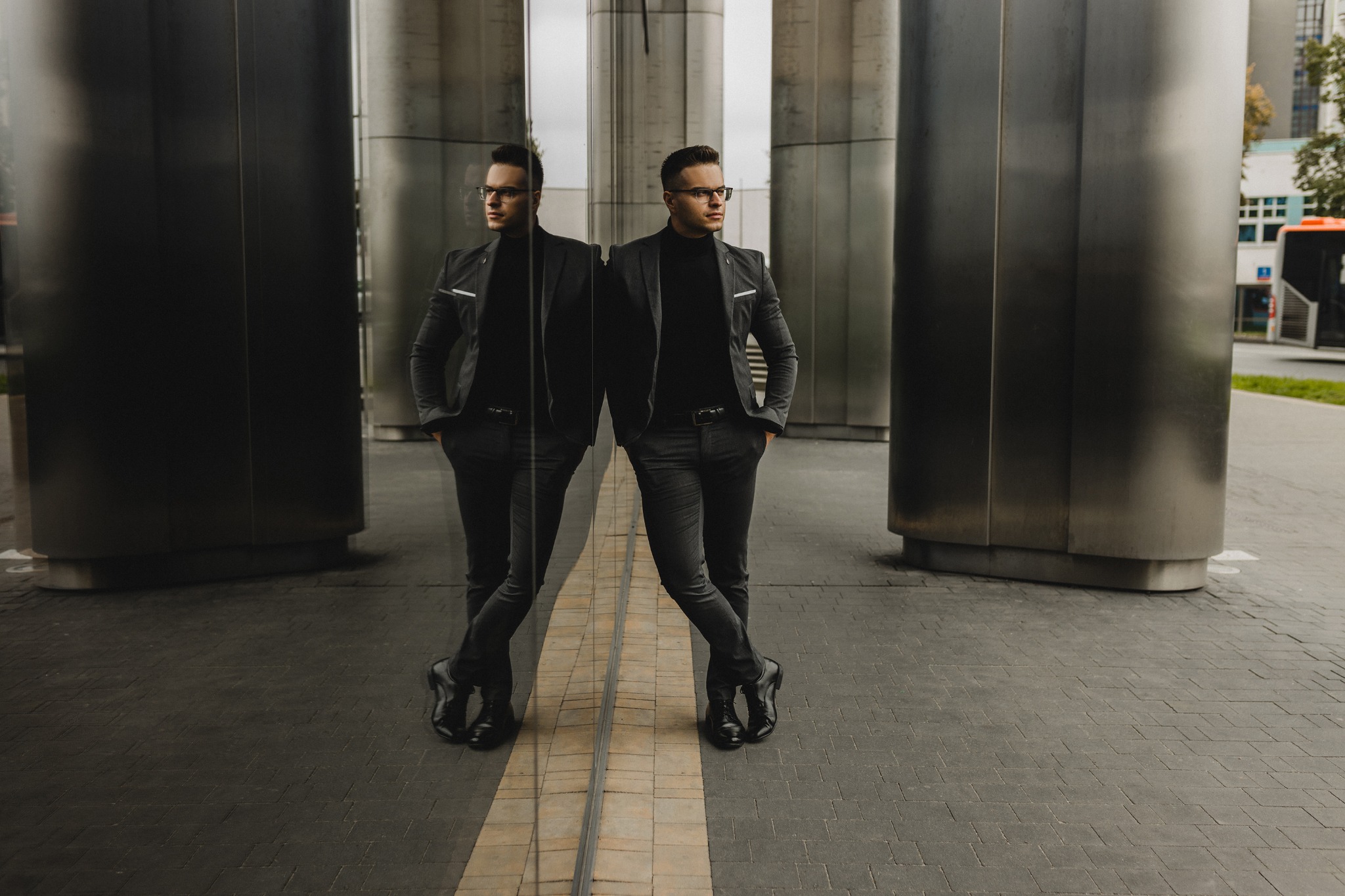 Mężczyzna w garniturze oparty o szklaną ścianę budynku, widoczne odbicie. Ujęcie z dołu, na zewnątrz, w tle fragment ulicy z autobusem. Neutralna kolorystyka.