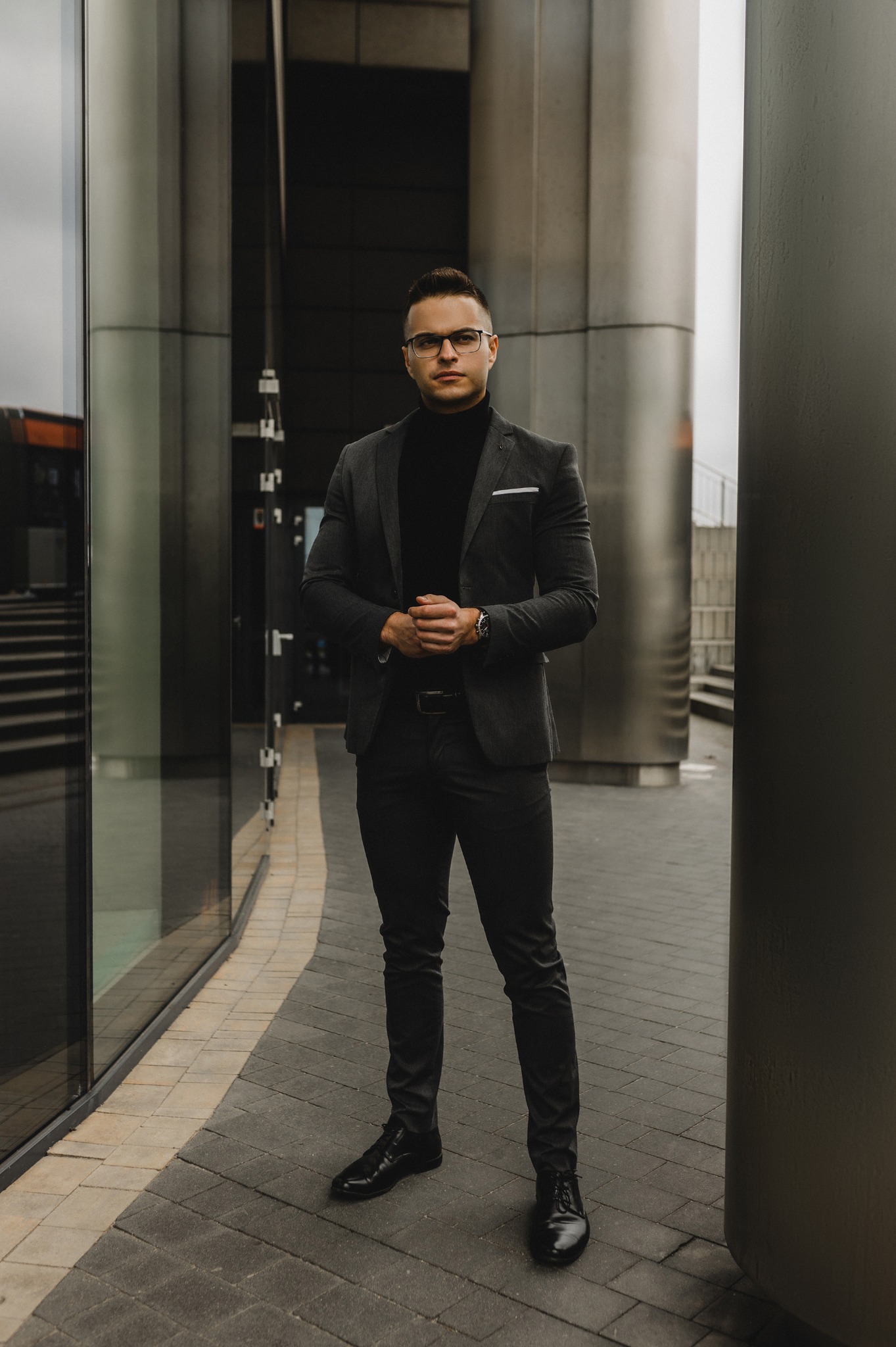 Mężczyzna w garniturze, czarnym golfie i okularach stoi na zewnątrz nowoczesnego budynku z szklaną fasadą. Elegancki wygląd, profesjonalny wizerunek.