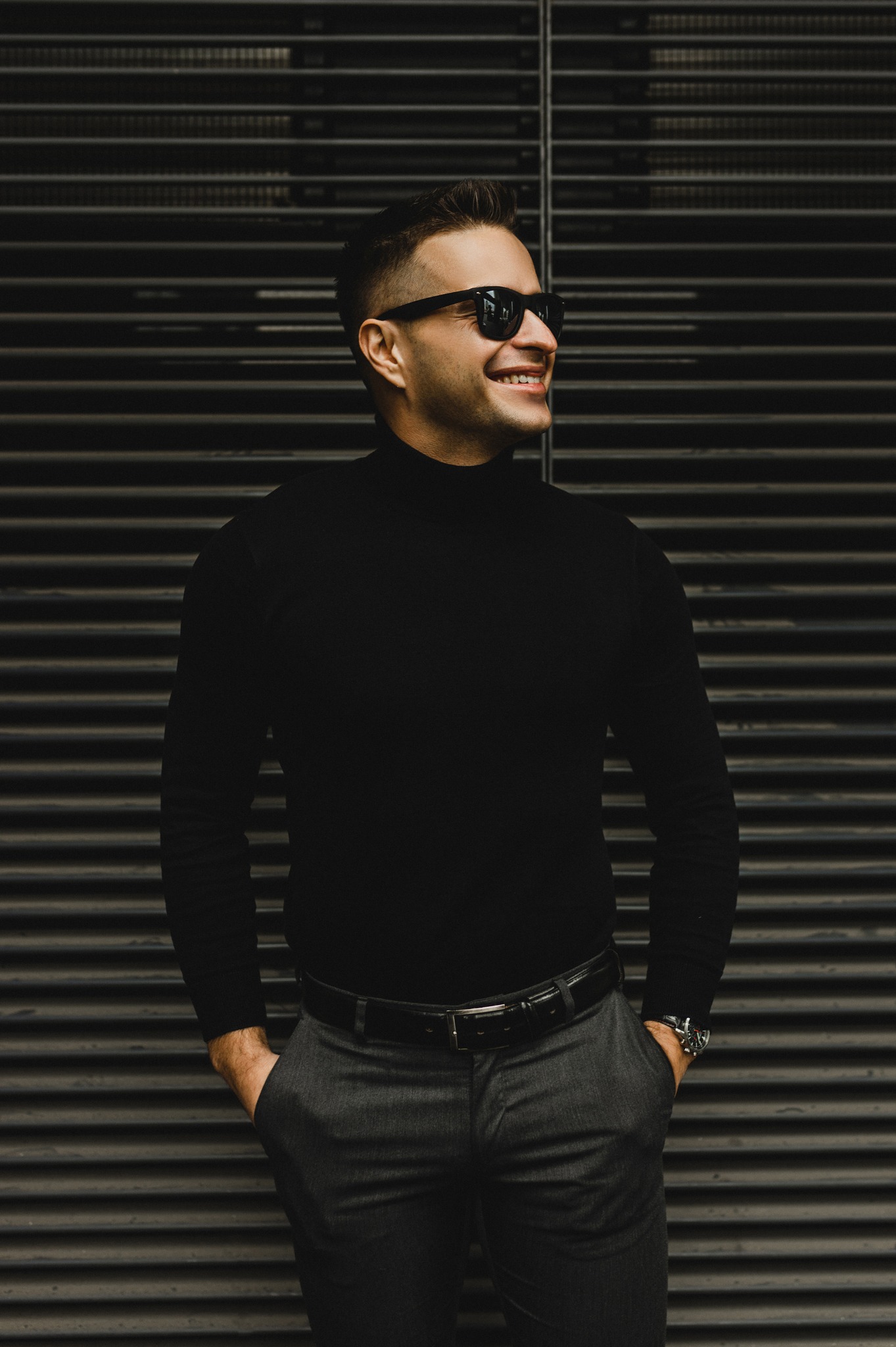Uśmiechnięty mężczyzna w czarnym golfie i okularach przeciwsłonecznych, z rękami w kieszeniach szarych spodni, na tle metalowej żaluzji. Widoczny zegarek na ręce.