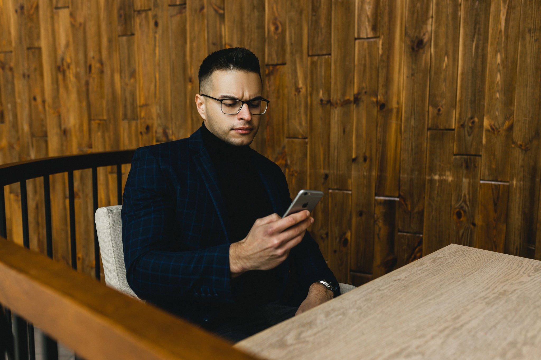 Mężczyzna w okularach i marynarce w kratę siedzi przy stole, przeglądając coś na smartfonie. W tle drewniana ściana i fragment balustrady.