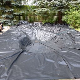 P&J Teramy Sp.Zo.o - Czarna folia rozłożona w wykopie pod oczko wodne, widoczne zmarszczki i fałdy, w tle drzewa iglaste i drewniany płot.