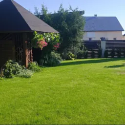 Zadbany trawnik z drewnianą altaną ozdobioną kwiatami, widok na dom z ciemnym dachem i nowoczesnym ogrodzeniem.