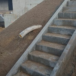 P&J Teramy Sp.Zo.o - Betonowe schody prowadzące na wzgórze z ziemią, z widoczną rurą drenażową i elementami konstrukcyjnymi w tle, pod błękitnym niebem.