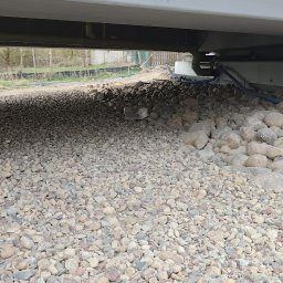 P&J Teramy Sp.Zo.o - Drenaż pod mostem: kamienie rzeczne i rura drenażowa. Widok na teren z zielenią w tle. Prace ziemne w toku.