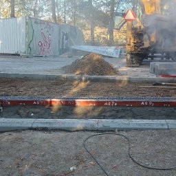 P&J Teramy Sp.Zo.o - Montaż odwodnienia liniowego z rurami oznaczonymi numerami, w tle koparka i hałda ziemi. Widoczny fragment kostki brukowej i zielony kask.