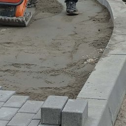 Jarosław Bełdycki - Układanie kostki brukowej: zagęszczarka wibruje piasek pod kostkę, widoczne krawężniki i częściowo ułożona kostka. Prace brukarskie w toku.