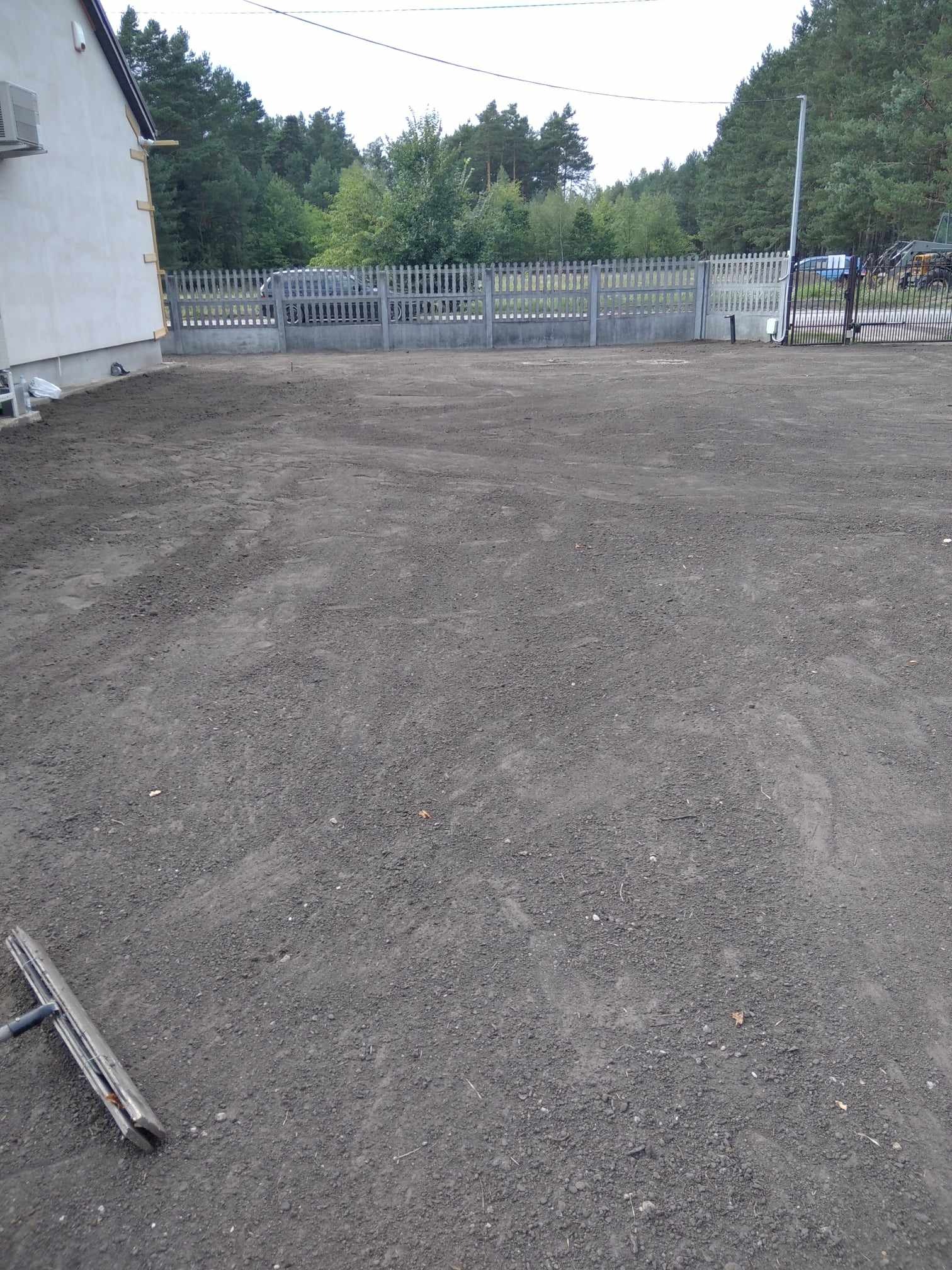 Przygotowany teren pod trawnik z widocznym narzędziem do wyrównywania, ogrodzeniem betonowym i fragmentem budynku z klimatyzatorem na ścianie.