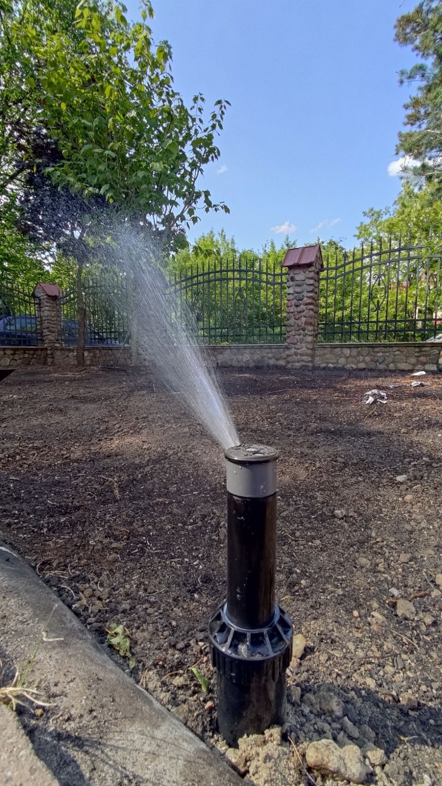 Automatyczny zraszacz ogrodowy w trakcie pracy, rozpylający wodę na świeżo przygotowaną ziemię, w tle widoczny kamienny murek z metalowym ogrodzeniem i zieleń drzew.