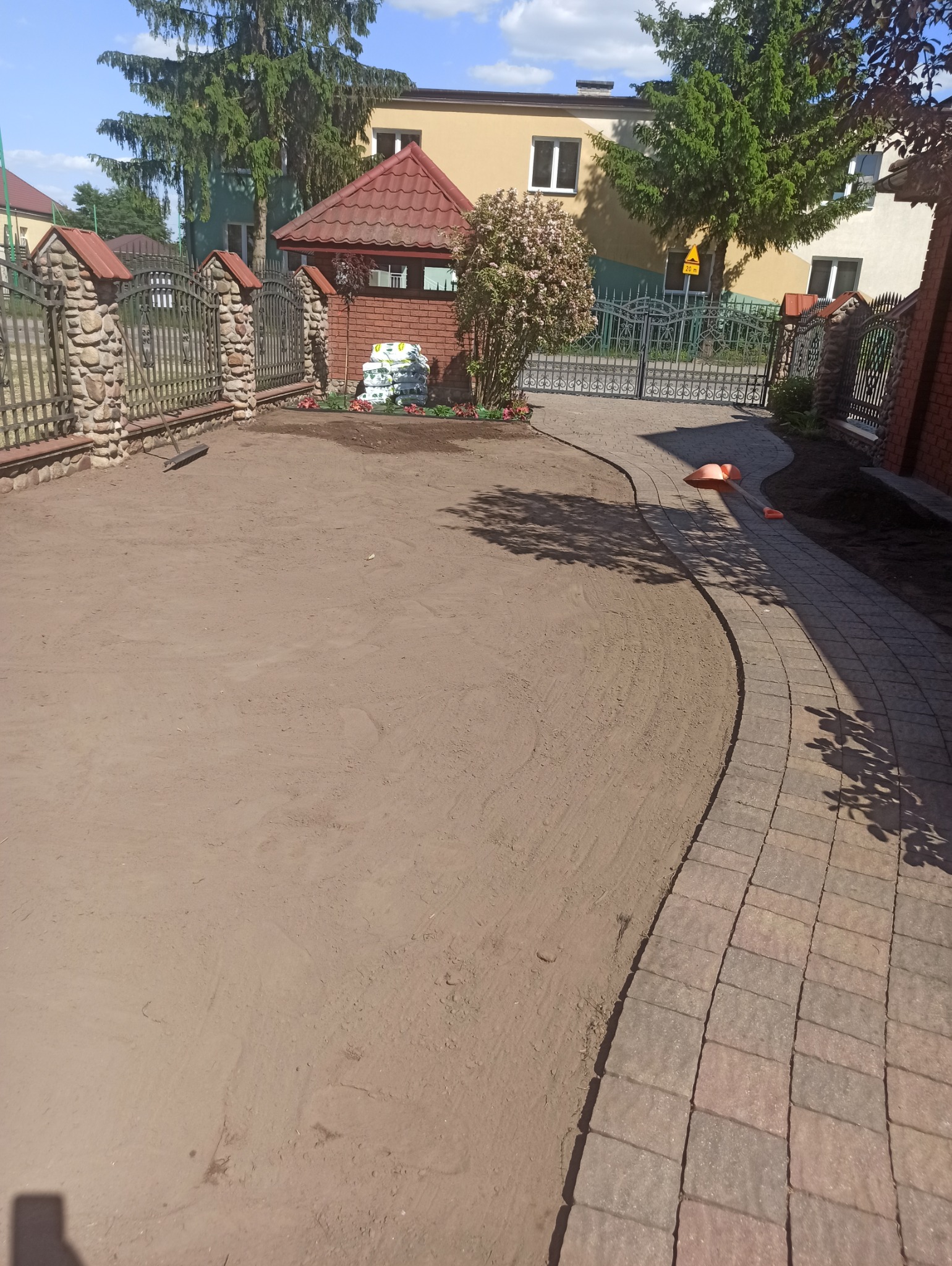 Przygotowany teren pod trawnik z widocznymi śladami po bronowaniu, obok ułożony chodnik z kostki, w tle ogrodzenie z kamiennymi słupkami i roślinność.