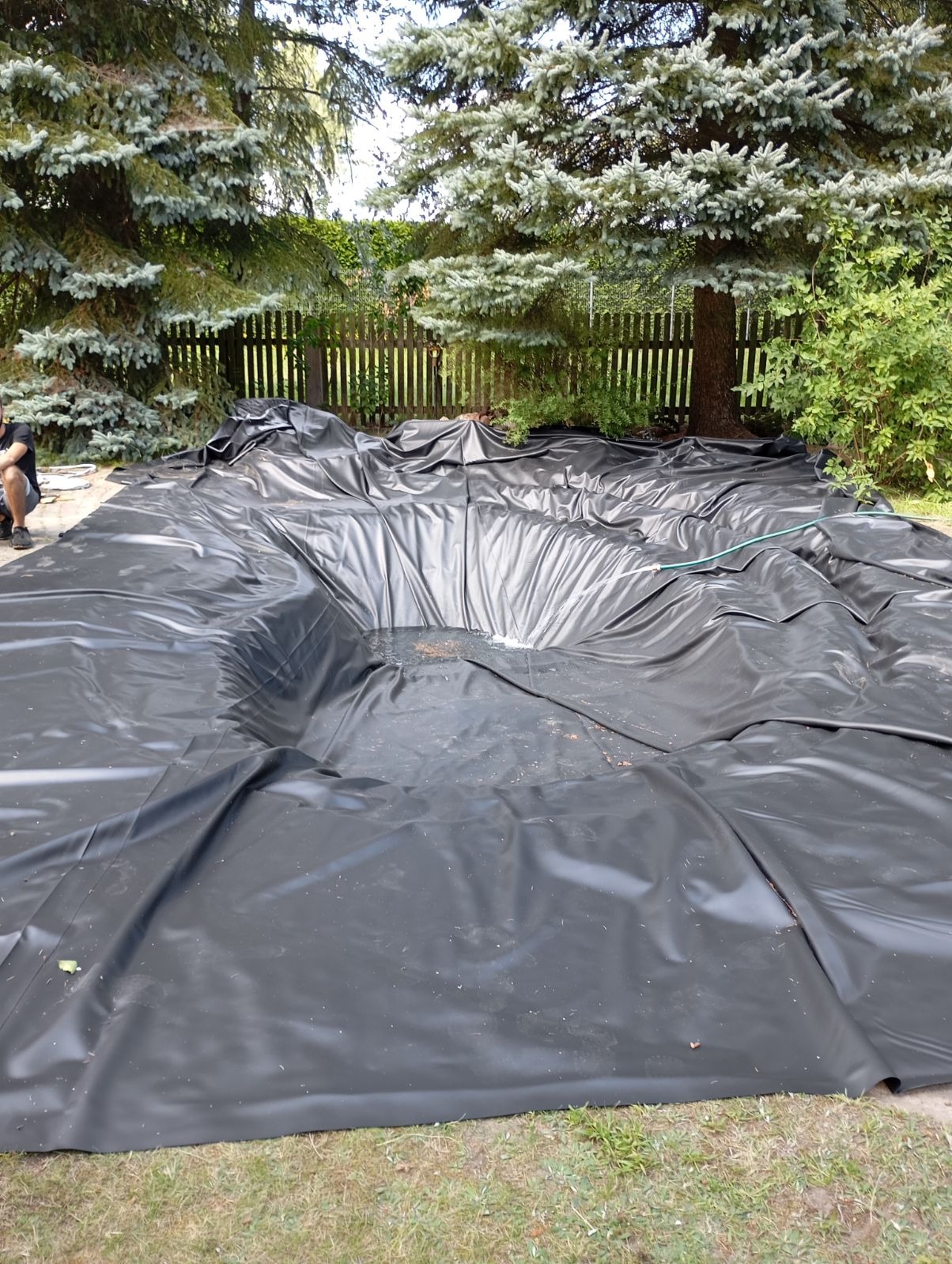 Czarna folia rozłożona w wykopie pod oczko wodne, widoczne zmarszczki i fałdy, w tle drzewa iglaste i drewniany płot.
