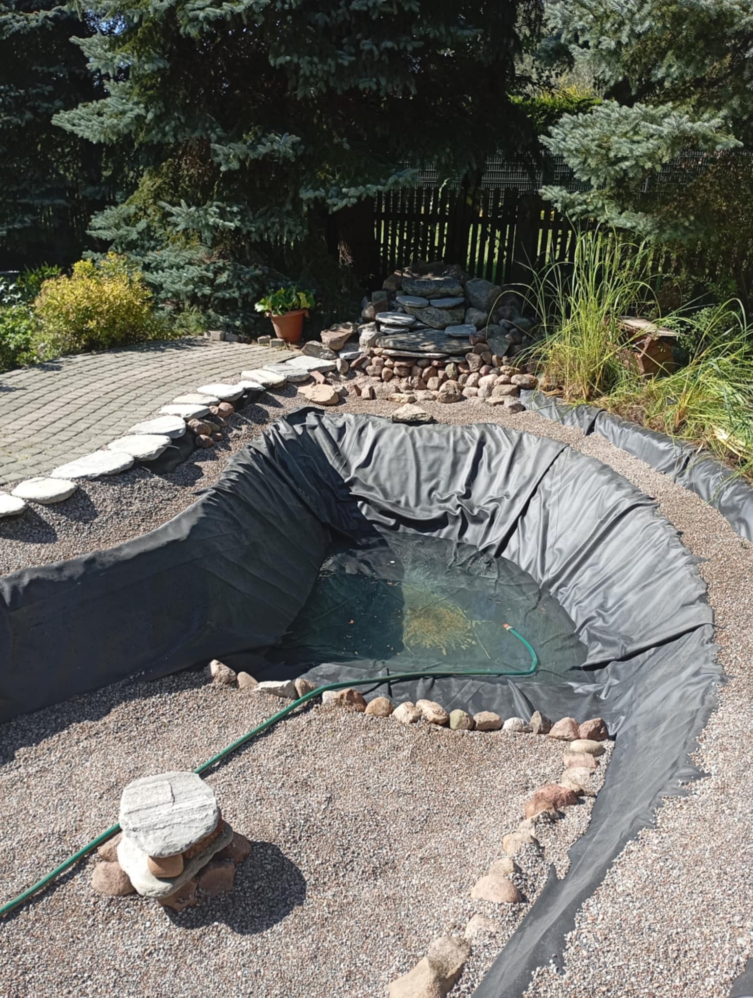 Wykopany dół wyłożony czarną folią, przygotowany pod budowę oczka wodnego w ogrodzie, z widocznym kamiennym wodospadem w tle i zielonym wężem ogrodowym doprowadzającym wodę.