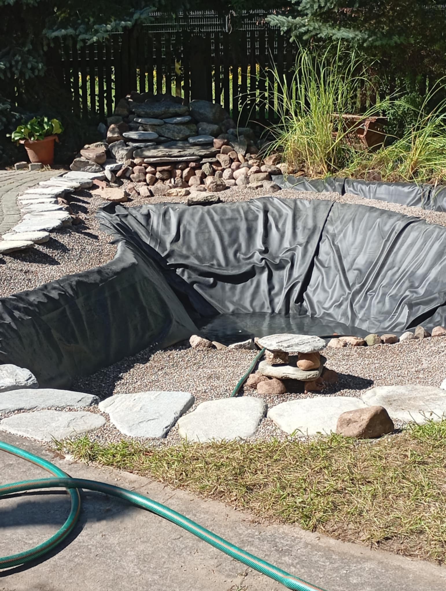 Budowa oczka wodnego w ogrodzie: wykop wyłożony czarną folią, kamienne obrzeże, kaskada z kamieni w tle, widoczny wąż ogrodowy.