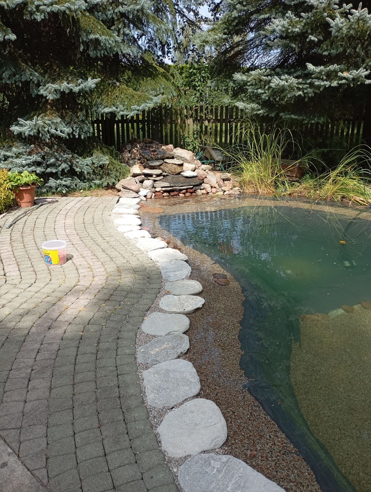 Oczko wodne z kaskadą wodną otoczone kamienną ścieżką i roślinnością, widoczne niebieskie świerki w tle, na pierwszym planie wiaderko z nawozem.