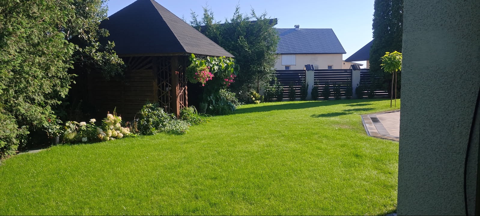 Zadbany trawnik z drewnianą altaną ozdobioną kwiatami, widok na dom z ciemnym dachem i nowoczesnym ogrodzeniem.
