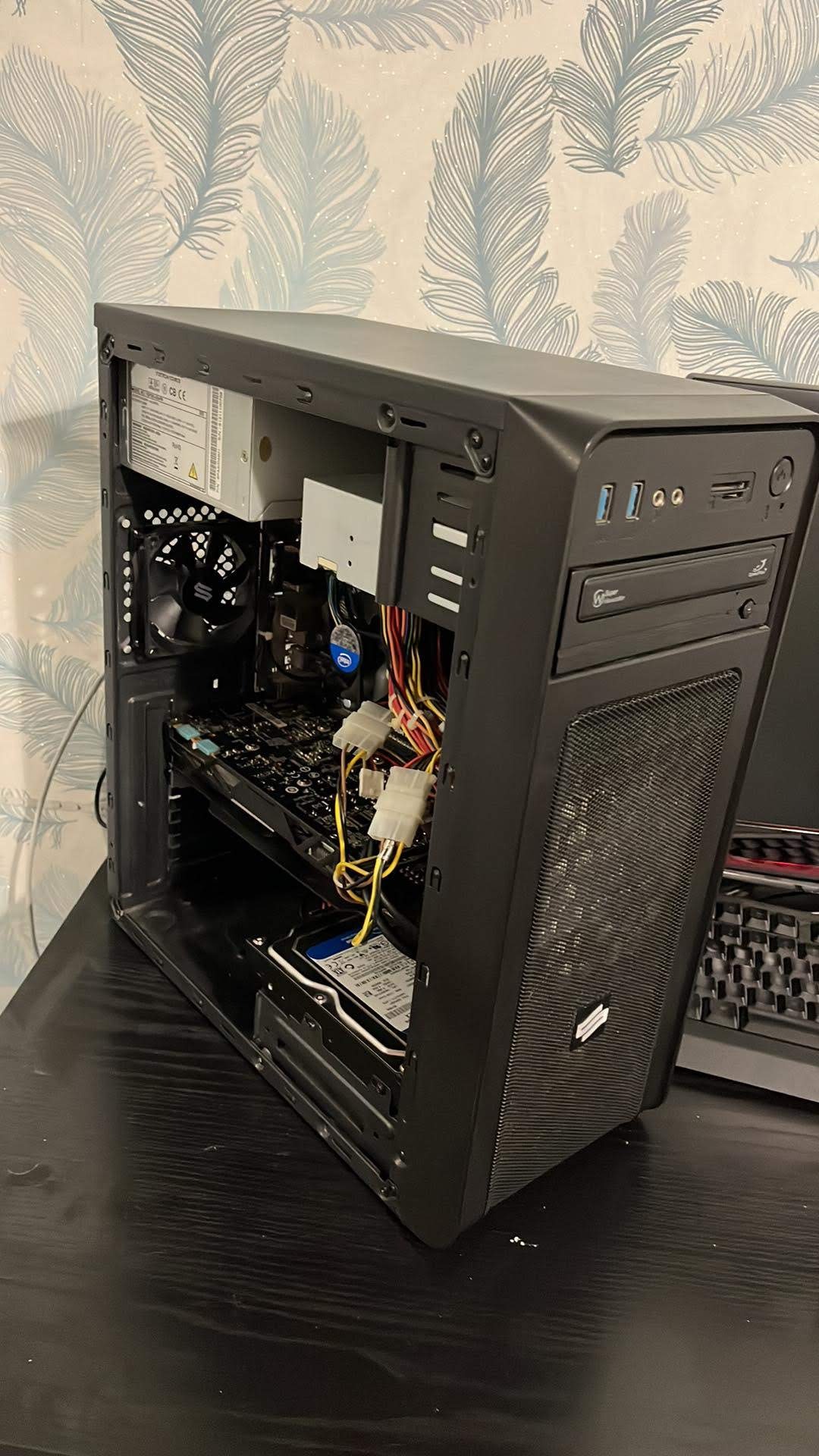 Otwarta obudowa komputera stacjonarnego z widocznymi podzespołami: karta graficzna, dysk twardy, zasilacz i plątanina kabli na tle ściany w piórka.
