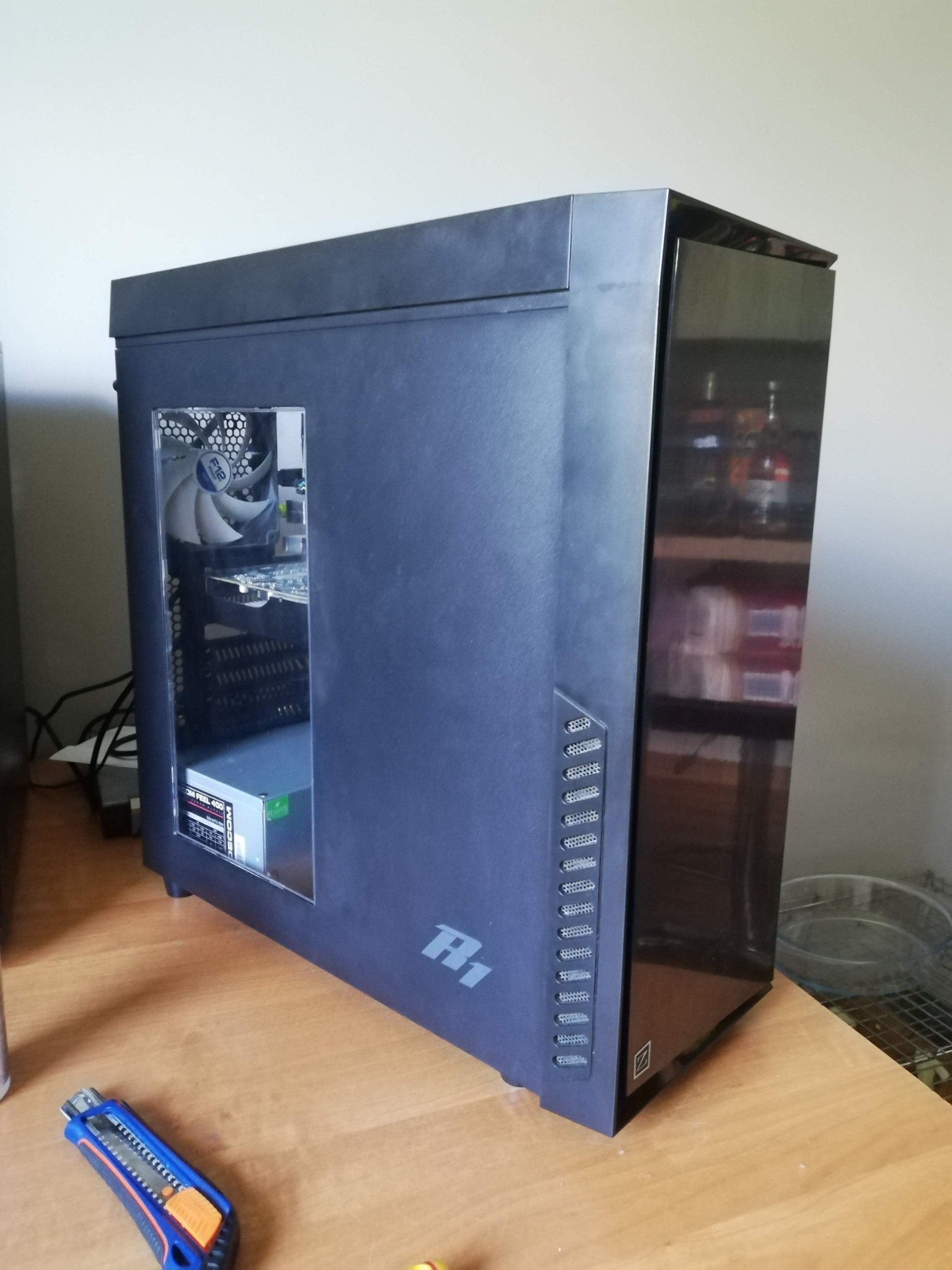 Komputer stacjonarny w czarnej obudowie z widocznym wnętrzem, w tym wentylator, karta graficzna i zasilacz, stoi na drewnianym biurku obok nożyka segmentowego.