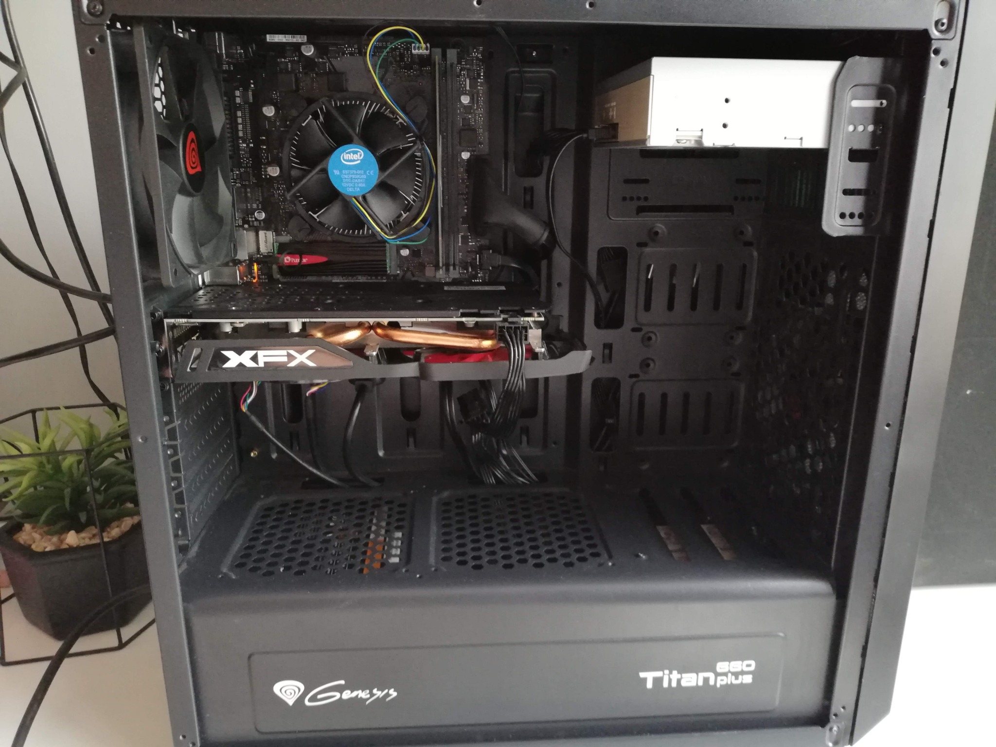 Wnętrze obudowy komputera stacjonarnego marki Genesis Titan Plus 660 z zamontowaną płytą główną Intel, kartą graficzną XFX i chłodzeniem procesora. Widoczny wentylator z logo Genesis w górnej...