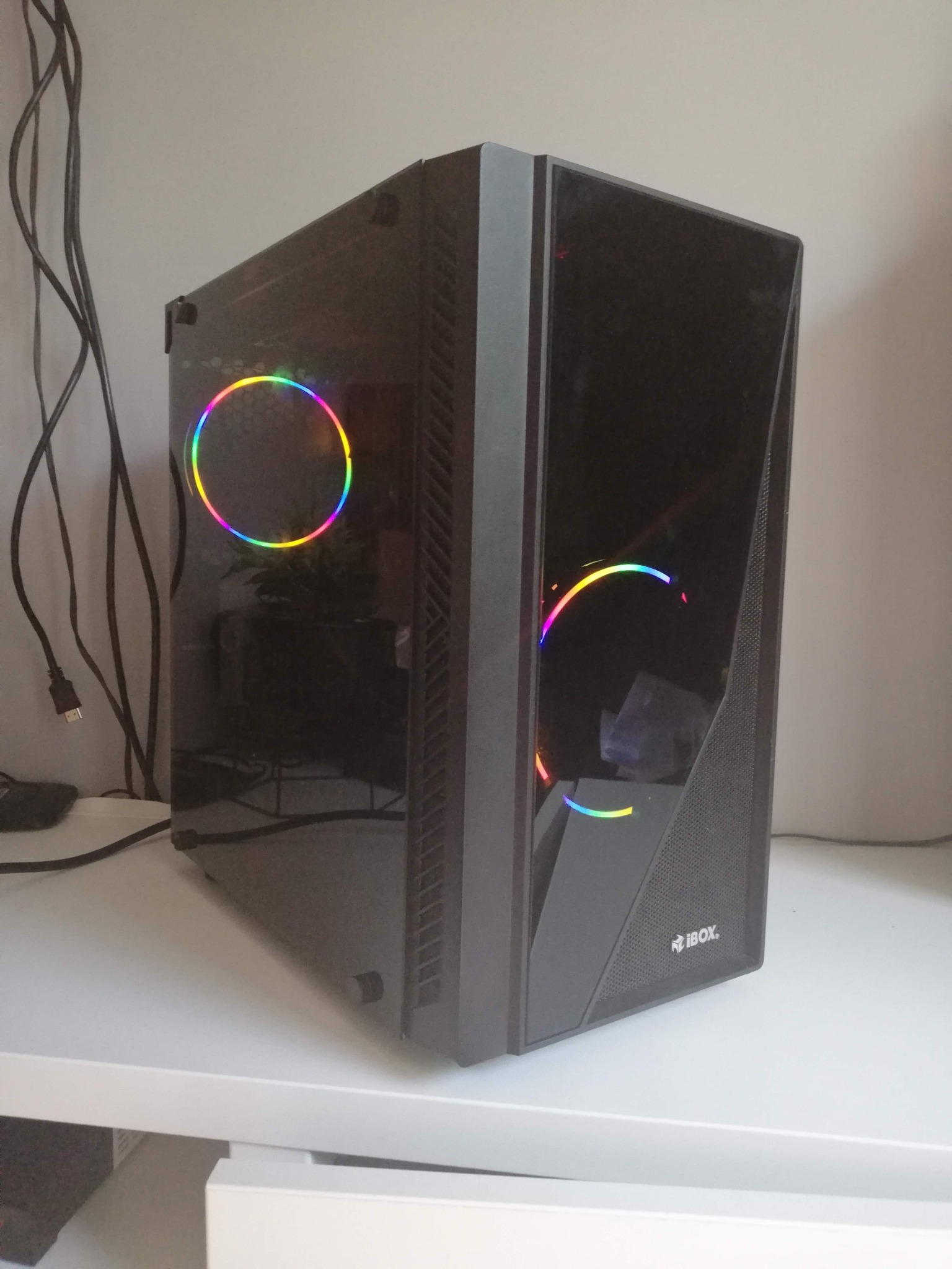 Czarna obudowa komputera stacjonarnego marki iBOX z podświetleniem RGB, widoczna na białym biurku, z kablami zasilającymi w tle.