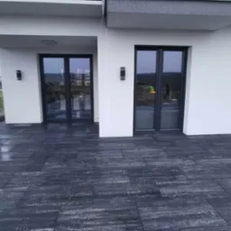 Nowoczesny taras wyłożony ciemnoszarą kostką brukową o nieregularnej fakturze, przylegający do białego budynku z grafitowymi ramami okiennymi i minimalistycznym oświetleniem zewnętrznym.