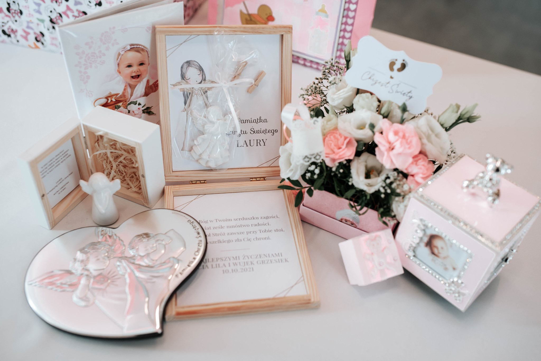 Pamiątki Chrztu Świętego Laury: drewniane ramki ze zdjęciami i sentencjami, srebrne serce z aniołami, bukiet kwiatów w różowym pudełku, upominki z motywem anioła, prezentacja na białym tle.