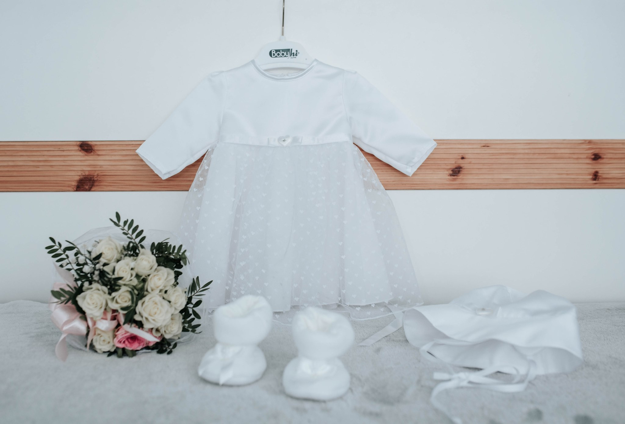Biała sukienka do chrztu zawieszona na wieszaku, obok bukiet białych róż z różową wstążką, białe buciki i czapeczka.