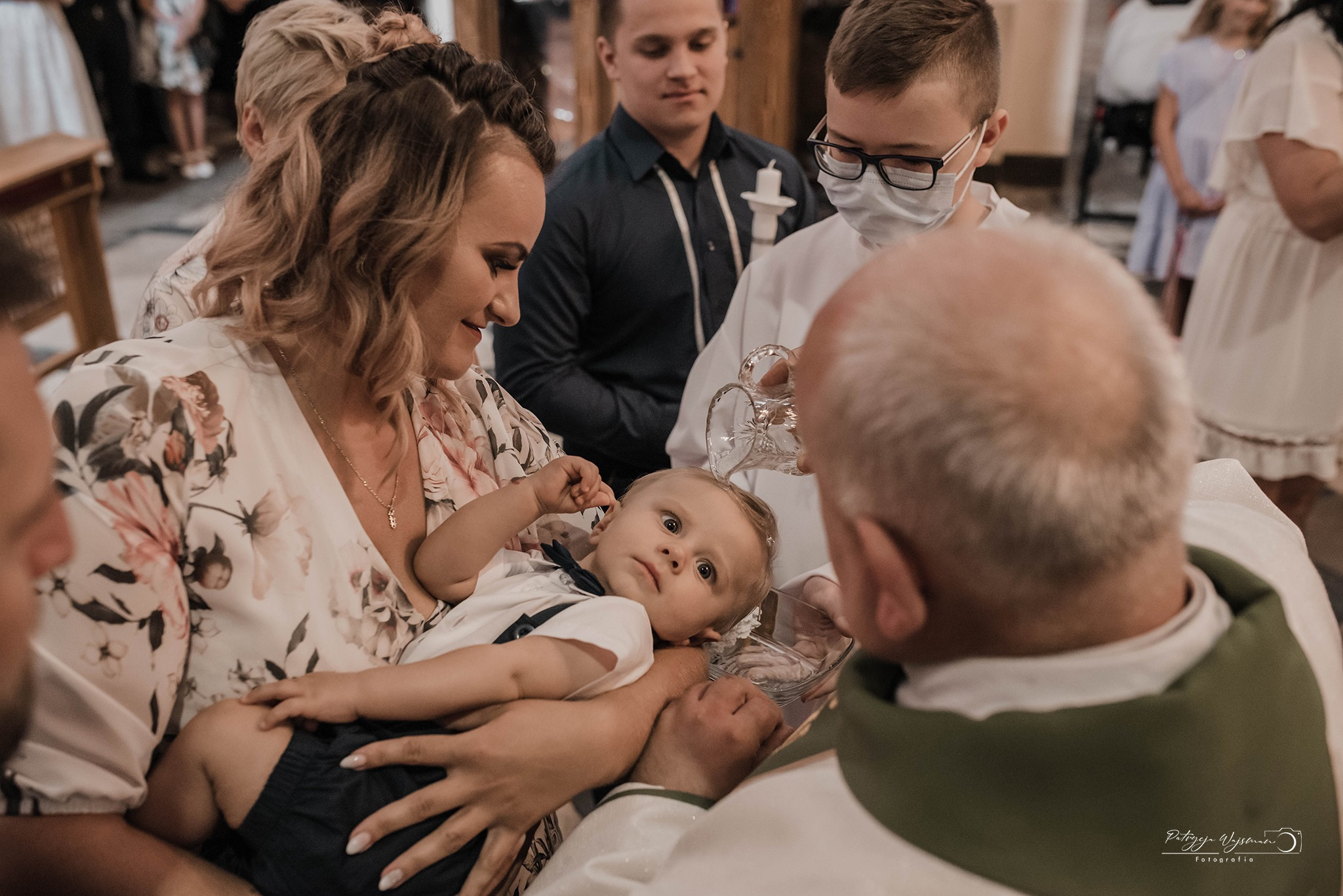 Matka trzyma niemowlę podczas chrztu, w tle ksiądz polewa głowę dziecka wodą z kryształowego naczynia, obecni goście.