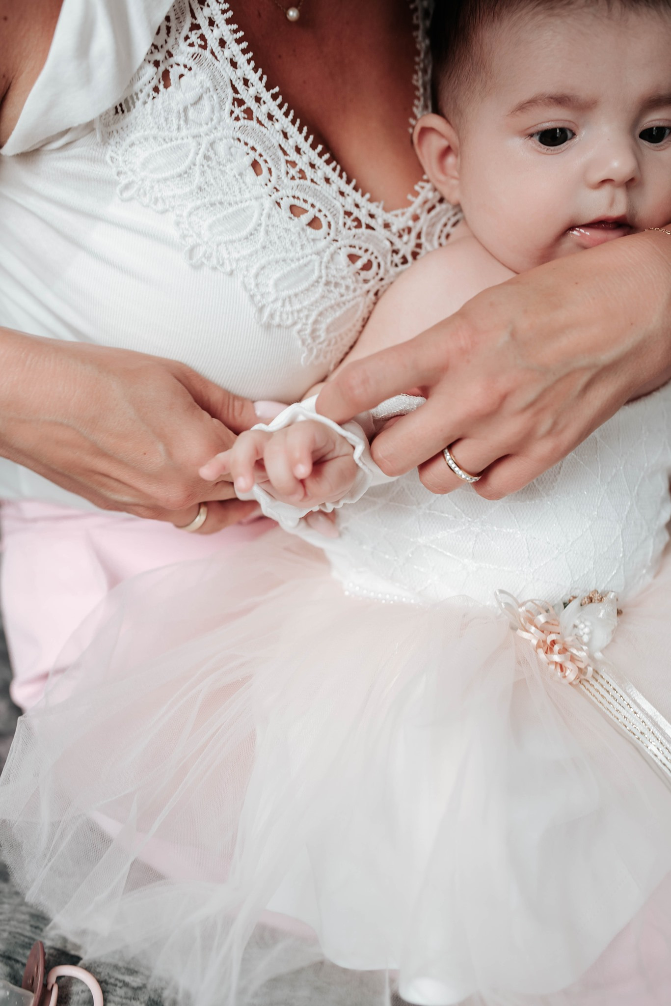 Mama trzyma na rękach niemowlę ubrane w białą sukienkę z tiulową spódniczką, zbliżenie na dłonie i małe rączki dziecka.