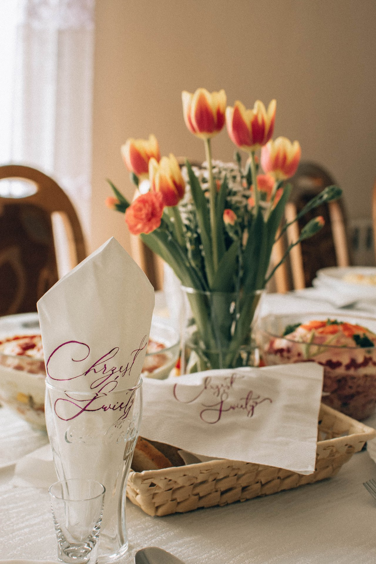 Uroczysty stół z dekoracjami na chrzest: tulipany w wazonie, sałatki w szklanych misach i serwetki z napisem 'Chrzest Święty' w ozdobnym piśmie.