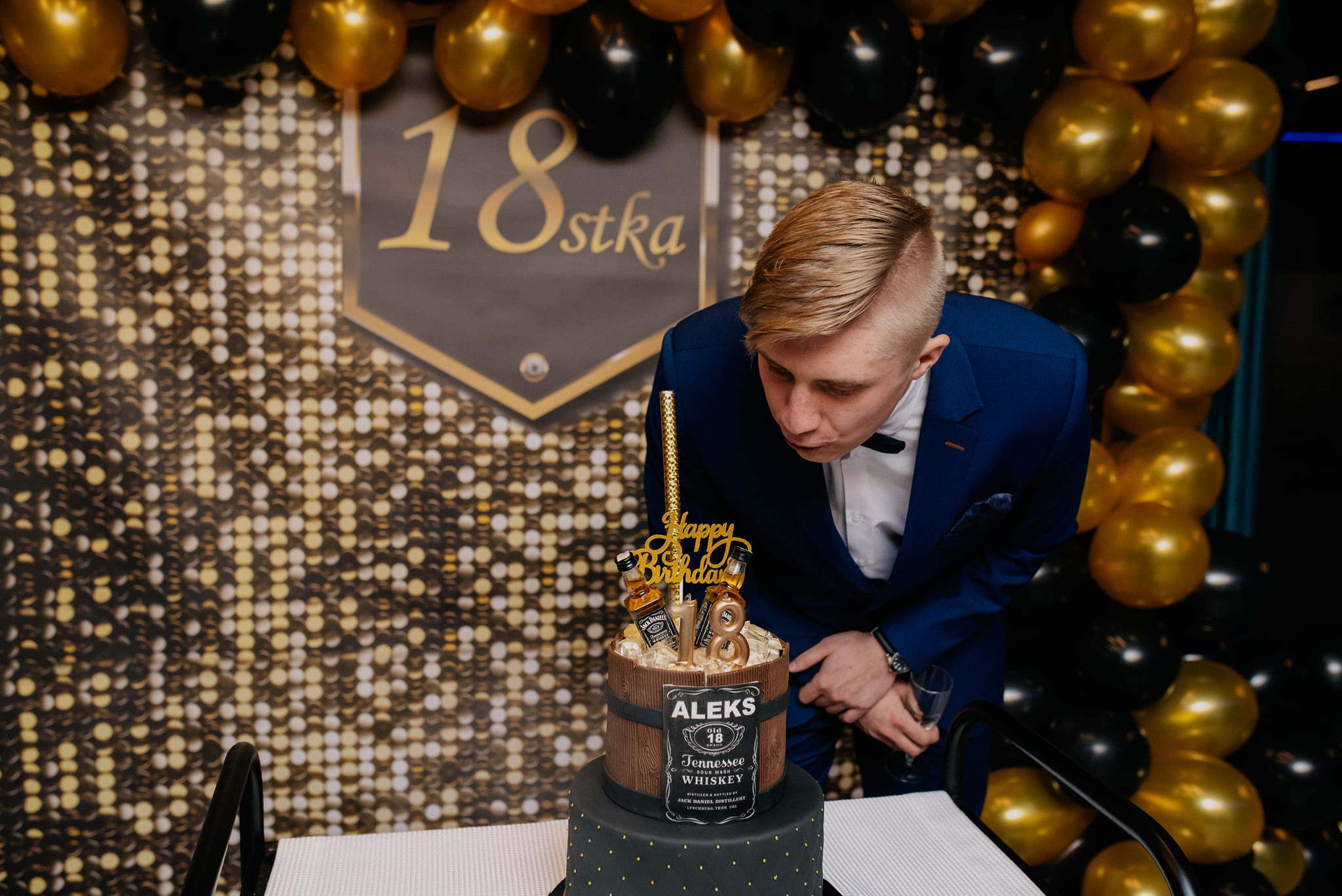 Młody mężczyzna w niebieskim garniturze i muszce dmucha świeczki na dwupoziomowym torcie urodzinowym z dekoracjami w stylu whiskey, na tle ściany z napisem '18stka' i balonów w kolorze złotym...