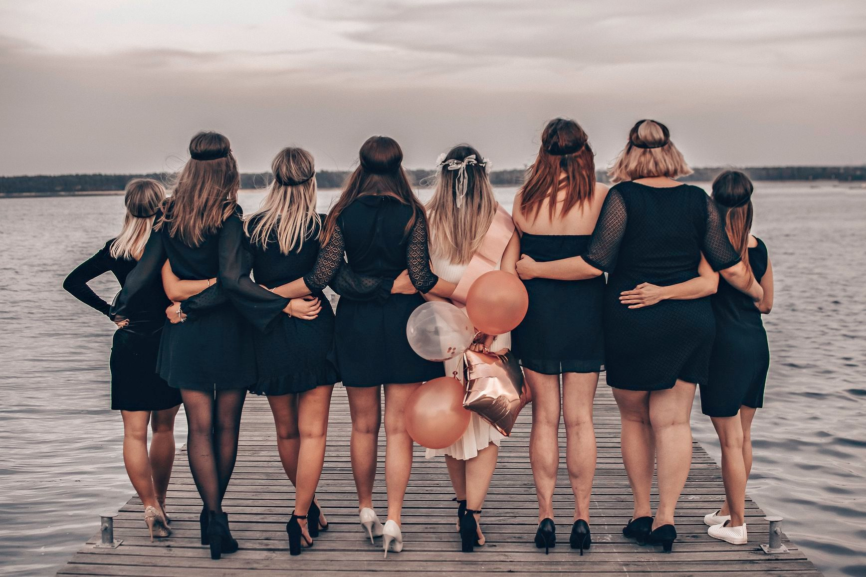 Grupa kobiet w czarnych sukienkach, obejmujących się na drewnianym molo z widokiem na wodę, jedna z kobiet trzyma balony, widok z tyłu.