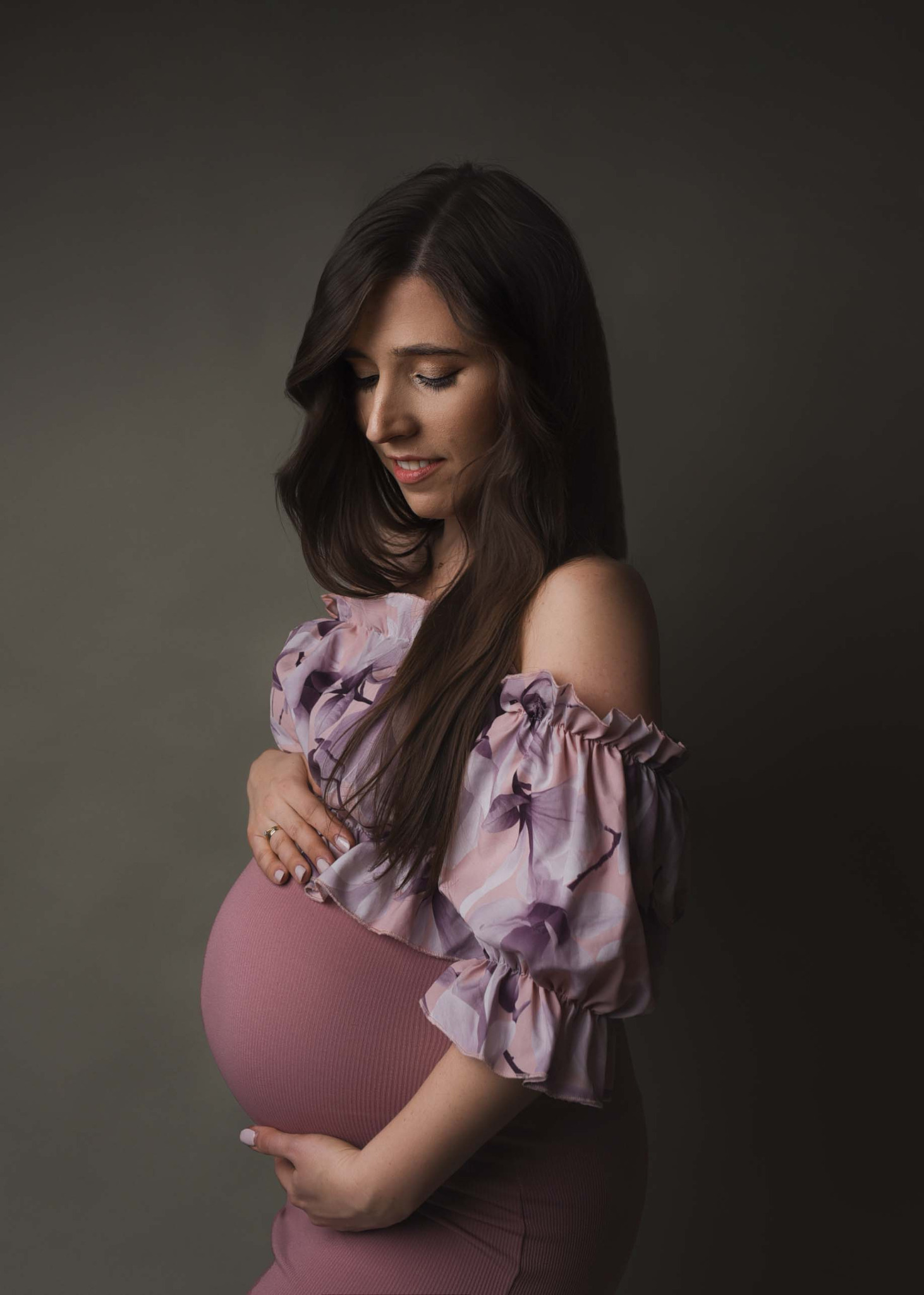Portret kobiety w ciąży w kwiecistej bluzce z odkrytymi ramionami i różowej sukience, spoglądającej w dół na swój brzuch, z pierścionkiem na palcu.