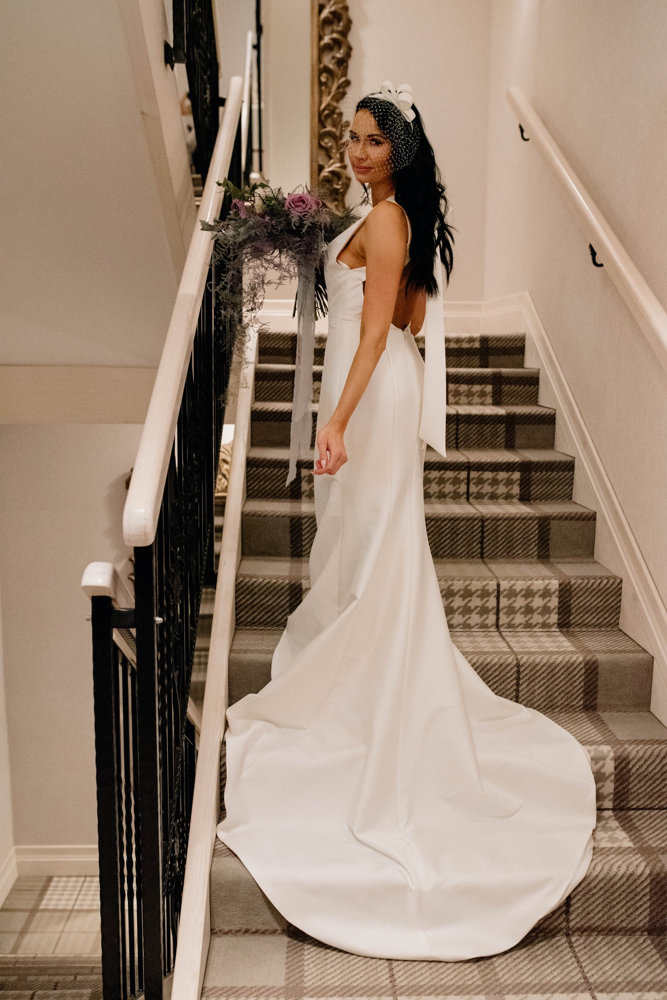 Panna młoda w białej sukni z odkrytymi plecami i bukietem, pozująca na schodach z wzorzystą wykładziną.