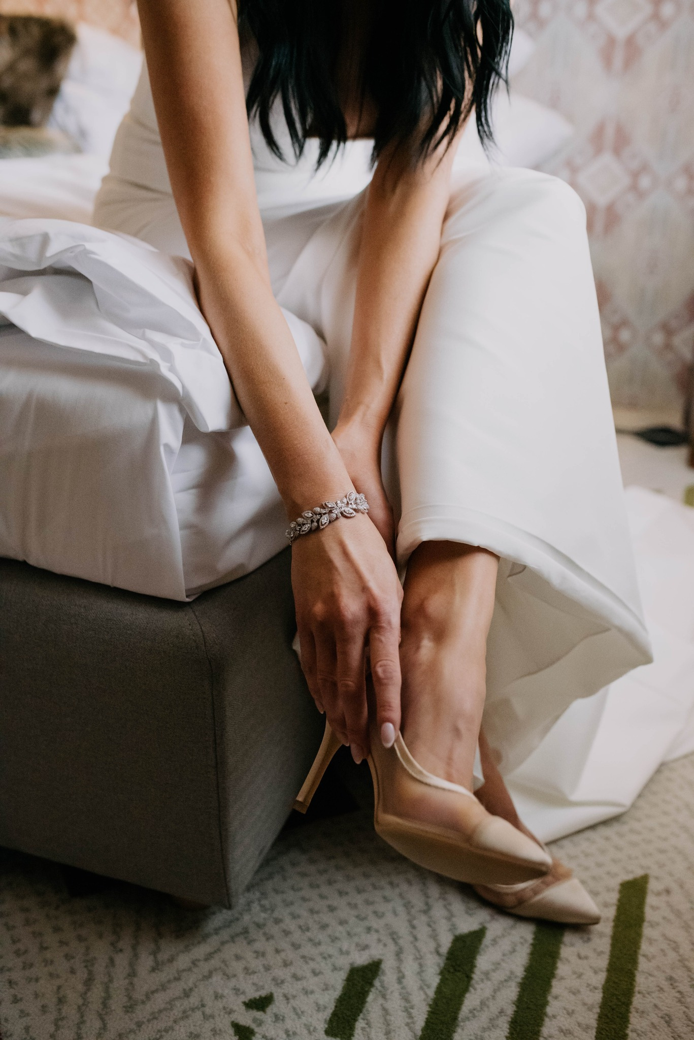 Panna młoda w białej sukni siedzi na łóżku, zapinając beżowy szpilkę, na nadgarstku błyszcząca bransoletka.