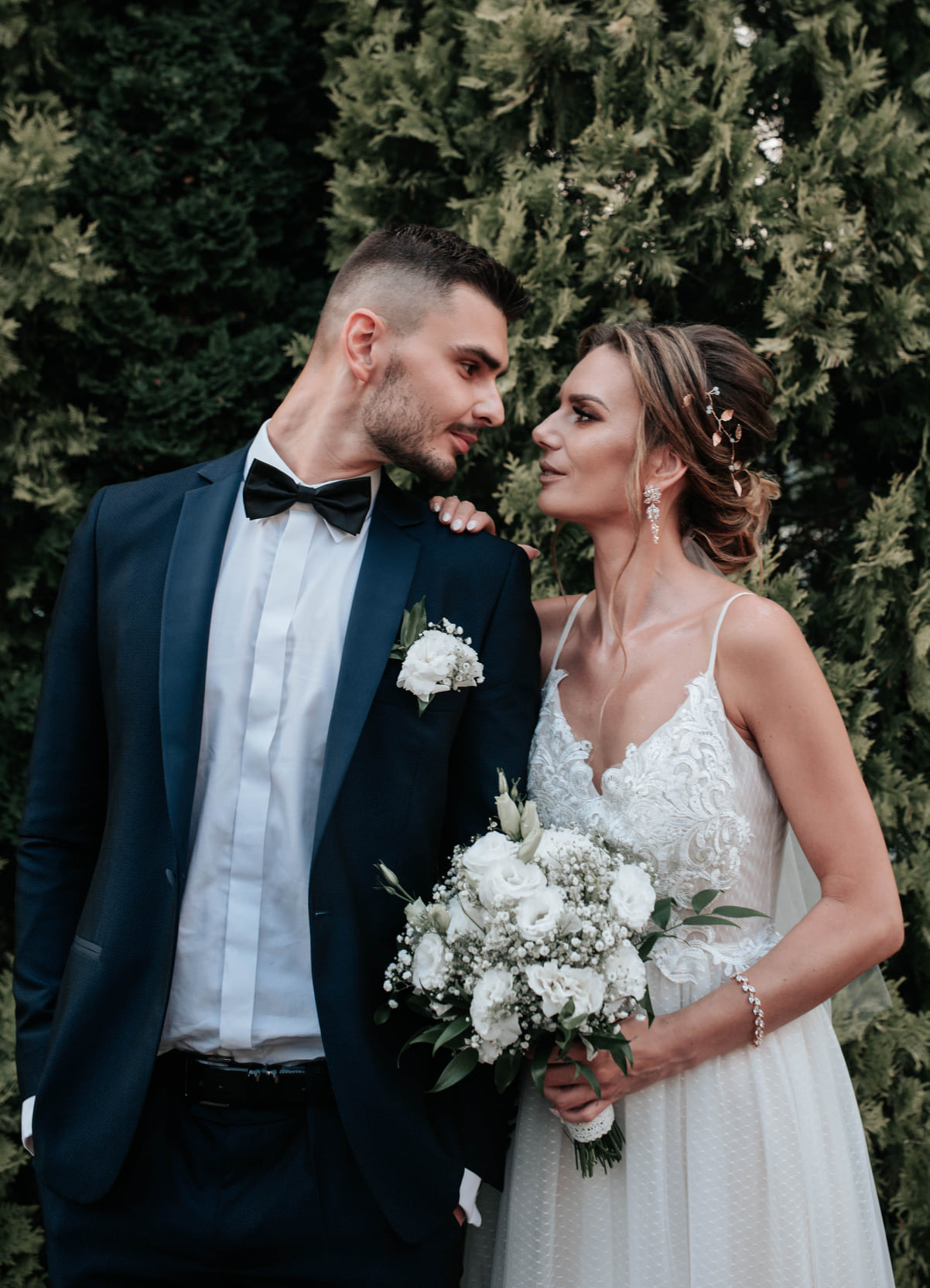 Elegancko ubrana para młoda, on w ciemnym garniturze z muchą, ona w białej sukni z koronkowym dekoltem i bukietem białych kwiatów, spoglądają na siebie na tle ciemnozielonych krzewów.
