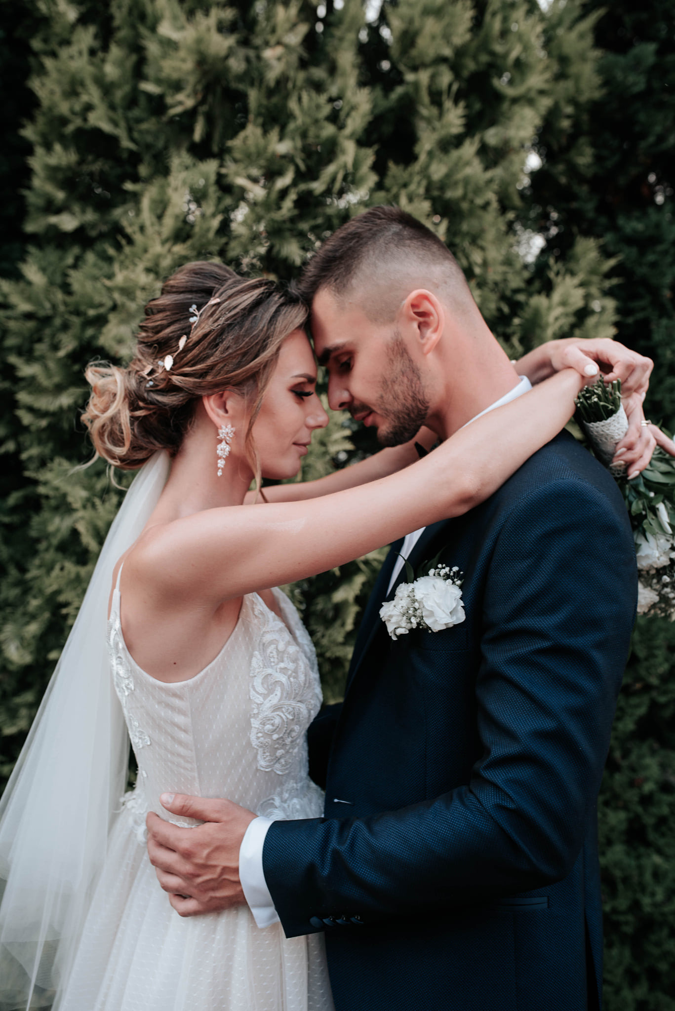 Para młoda, delikatny uścisk, panna młoda w sukni z koronkowymi aplikacjami, elegancki bukiet, ciemny garnitur pana młodego, zielone tło z krzewów.