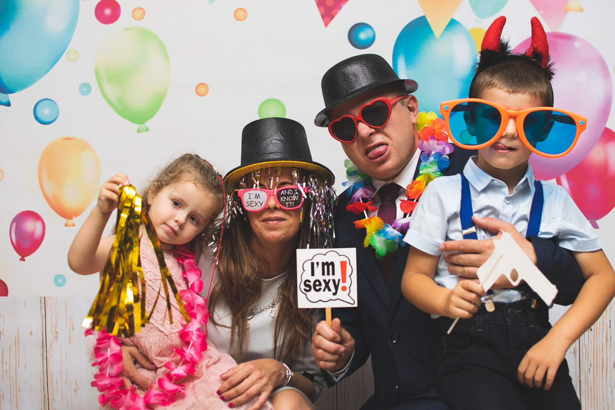 Rodzina pozuje do zdjęcia z rekwizytami: kapelusze, okulary w kształcie serc, hawajskie naszyjniki, papierowa broń i tabliczki z napisami przed tłem z balonami.