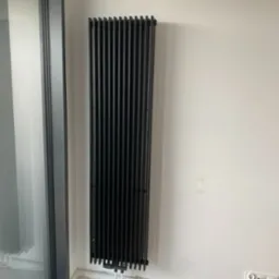 Czarny, pionowy grzejnik panelowy zamontowany na białej ścianie, widoczne podłączenia rur, obok przesuwne drzwi balkonowe.