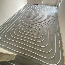 ZAKŁAD INSTALATORSTWA SANITARNEGO JAROSŁAW KOT - Białe rury ogrzewania podłogowego ułożone spiralnie na szarej macie izolacyjnej z siatką, widok z góry, fragment buta pracownika w rogu kadru.