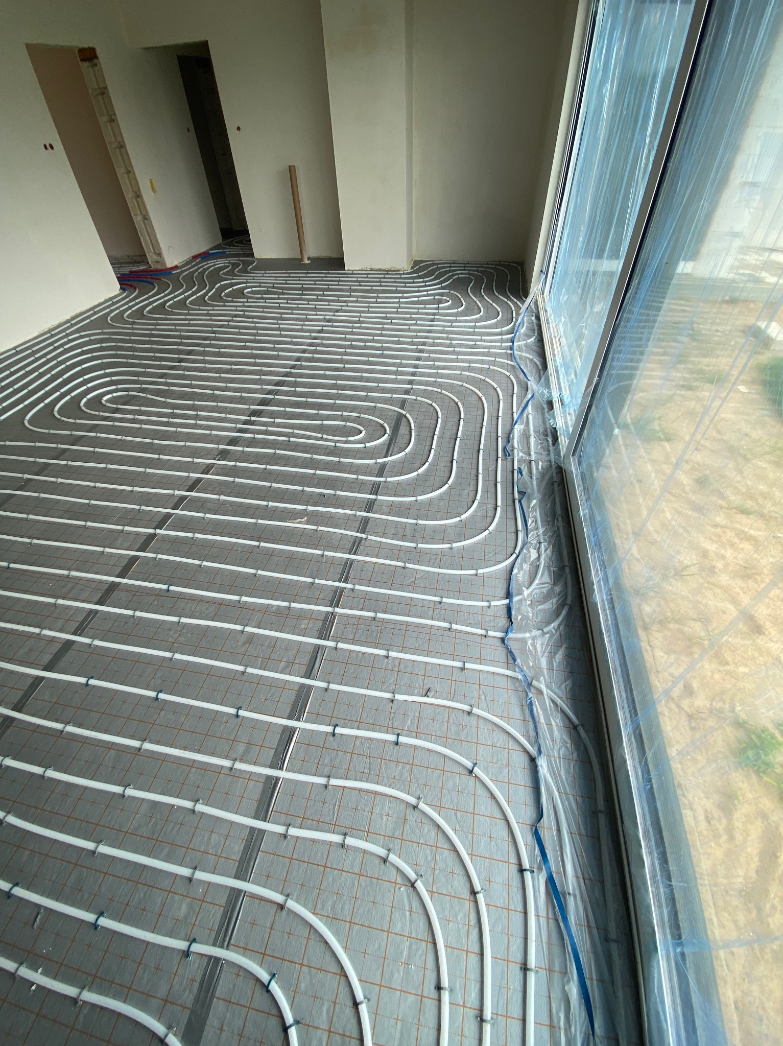 Białe rury ułożone w pętle na szarej macie z siatką, przygotowane do zalania w posadzce ogrzewania podłogowego, widok z góry w surowym wnętrzu z oknem zabezpieczonym folią.