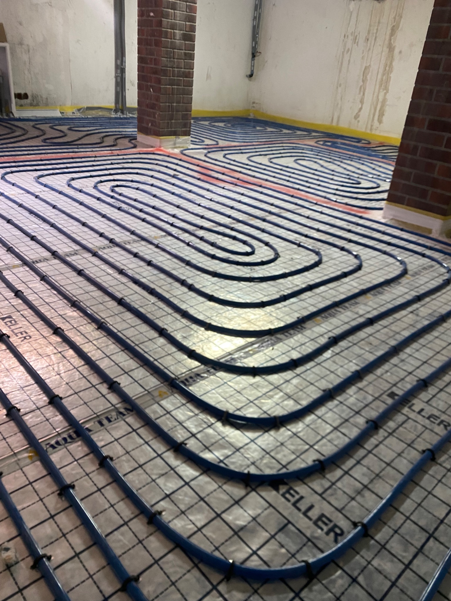 Równomiernie ułożone, niebieskie rury ogrzewania podłogowego na srebrnej siatce z widocznym napisem 'KELLER' i 'AQUA TEAM', z fragmentem kolumny z czerwonej cegły w tle, w surowym pomieszczeniu...