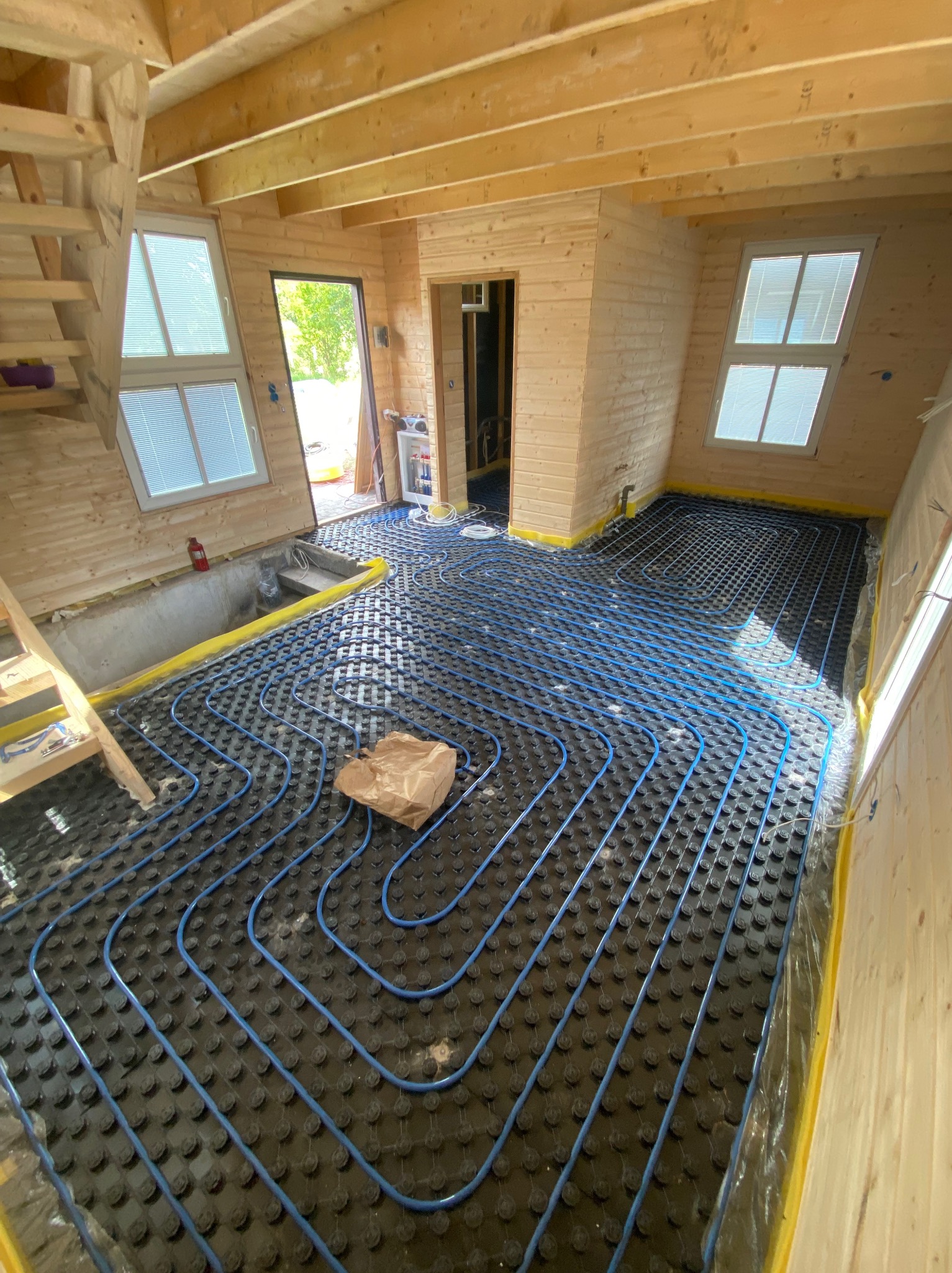 Instalacja ogrzewania podłogowego z niebieskimi rurami na czarnej macie w drewnianym domu w trakcie budowy, widoczne okna i schody.
