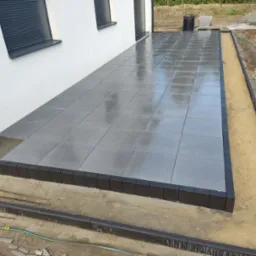 Nowoczesny taras z szarych płyt, obrzeża z ciemnej kostki. Widoczne przygotowania do dalszych prac ziemnych wokół tarasu. Świeżo ułożona nawierzchnia.