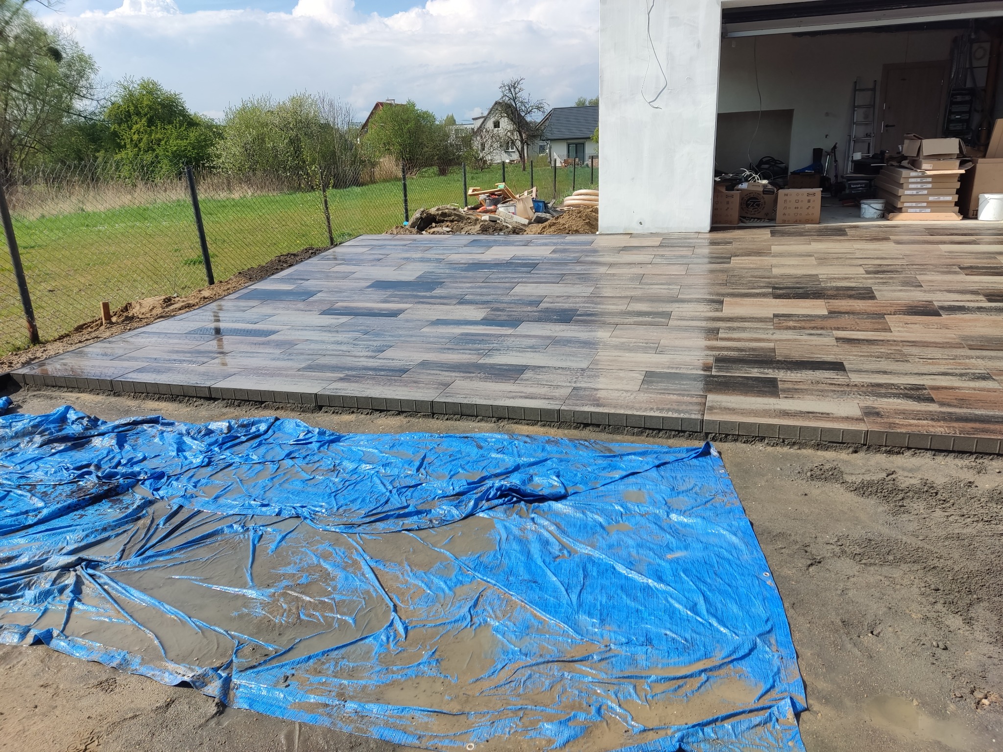 Ułożona kostka brukowa w odcieniach szarości i brązu, imitująca drewno, na posesji z garażem. Część kostki przykryta niebieską plandeką.