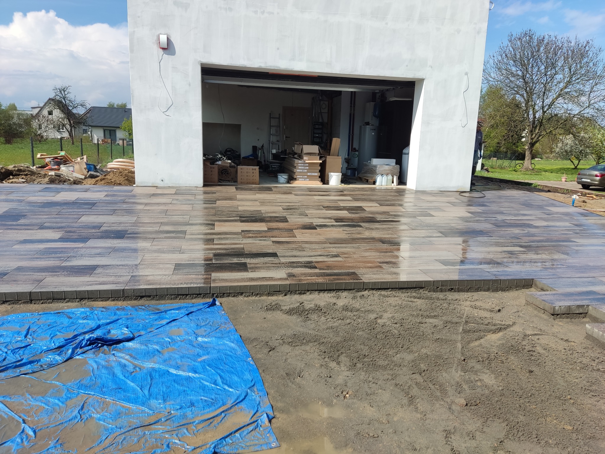Częściowo ułożona, błyszcząca kostka brukowa w odcieniach brązu i szarości, widoczna na podjeździe przed garażem. Prace w toku, widoczne narzędzia i materiały budowlane.