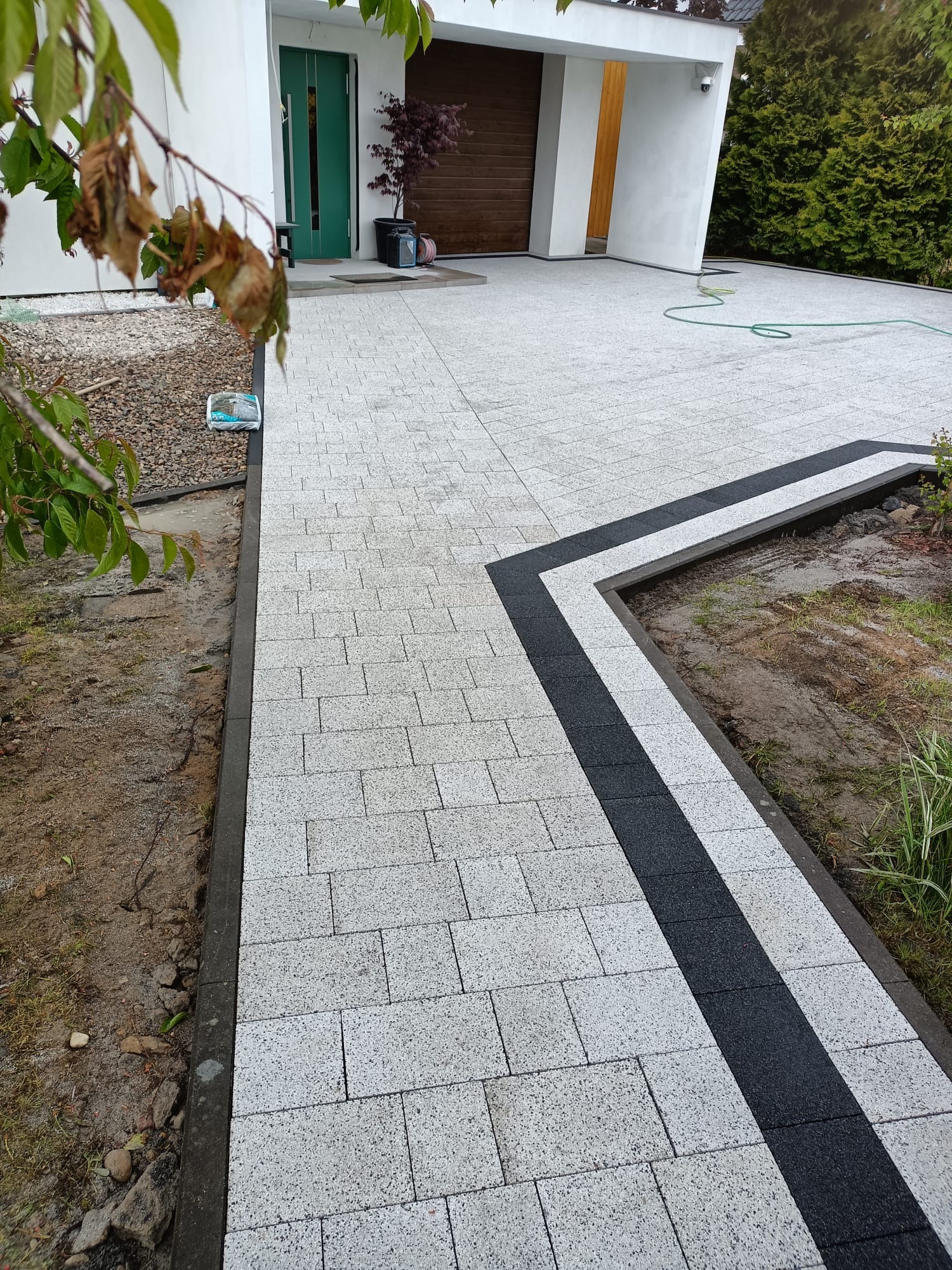 Jasnoszara kostka brukowa ułożona wzdłuż podjazdu, z czarnym pasem kontrastowym. Widoczny fragment ogrodu i domu z zielonymi drzwiami.