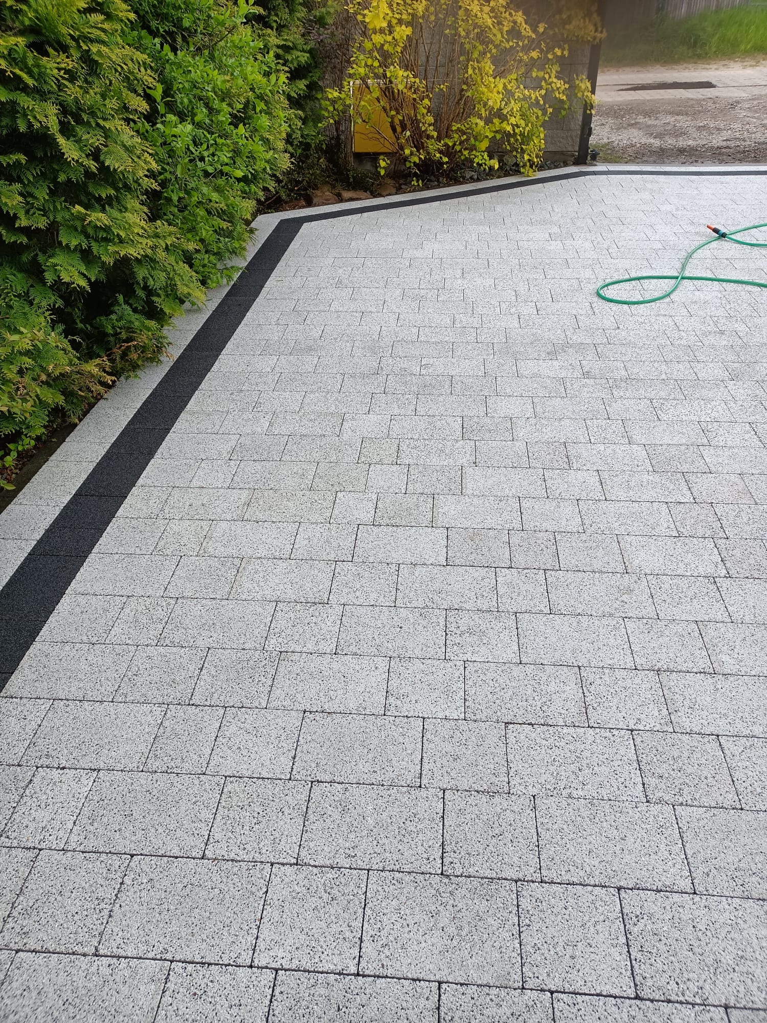 Ułożona jasnoszara kostka brukowa z ciemnoszarym obramowaniem, widok z góry. W tle zieleń i fragment węża ogrodowego. Estetyczne wykończenie podjazdu.