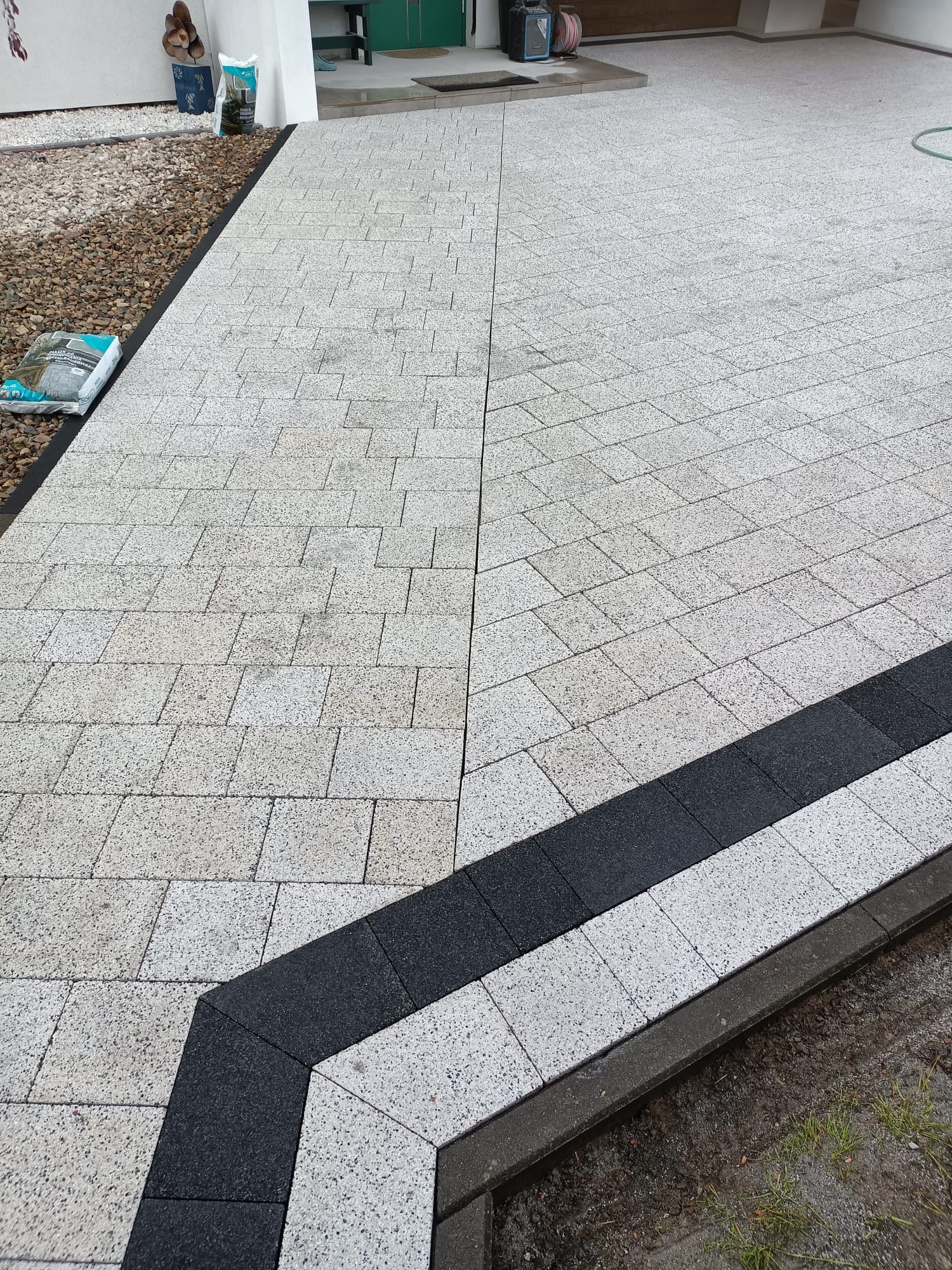 Jasnoszara kostka brukowa ułożona w geometryczny wzór z czarnymi obwódkami, widok z góry na podjazd. W tle widoczny fragment budynku.