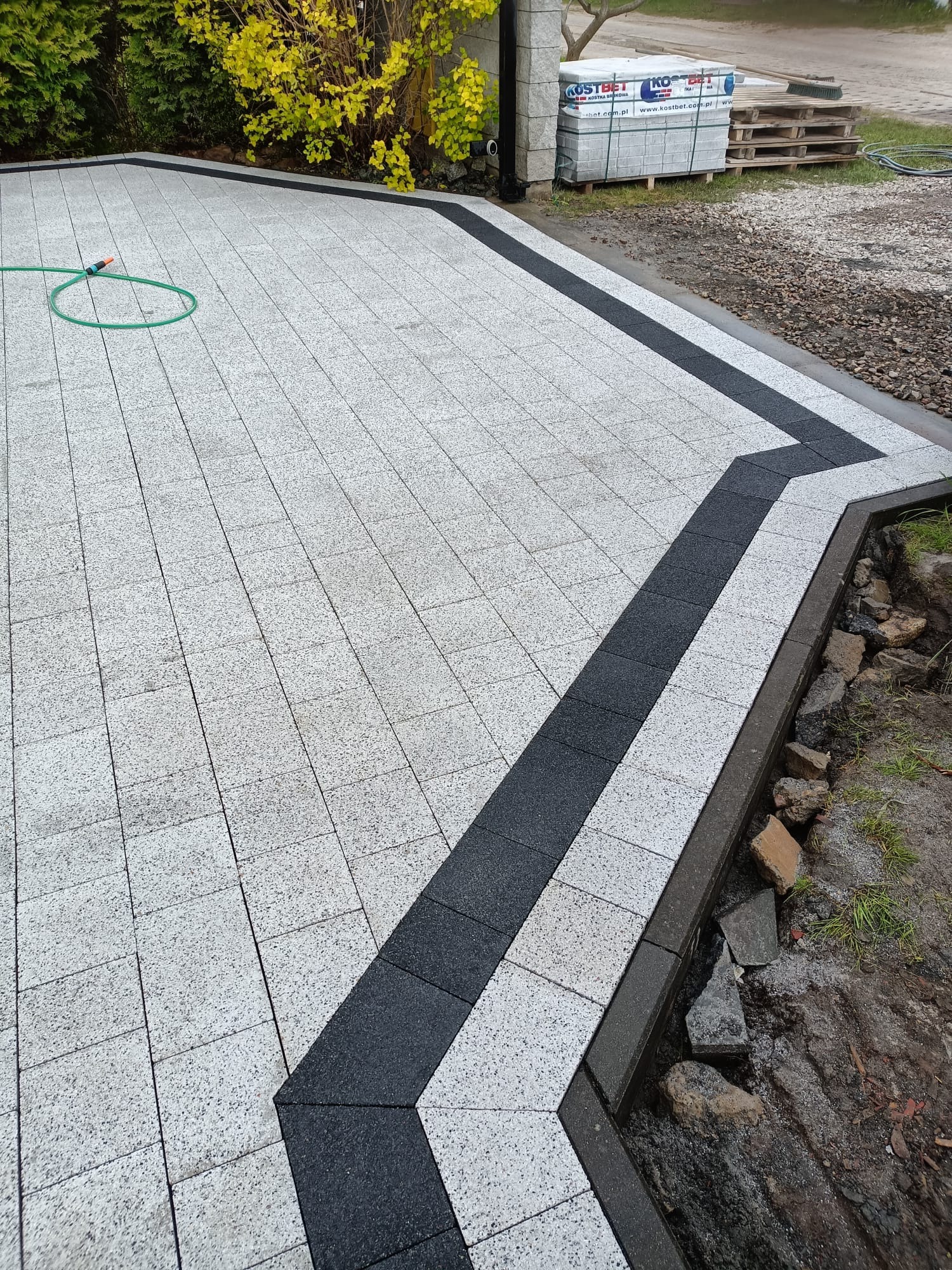 Ułożona kostka brukowa w jasnoszarym odcieniu z ciemnoszarym pasem, tworzącym ozdobny wzór. W tle widoczne palety z kostką i fragment roślinności.