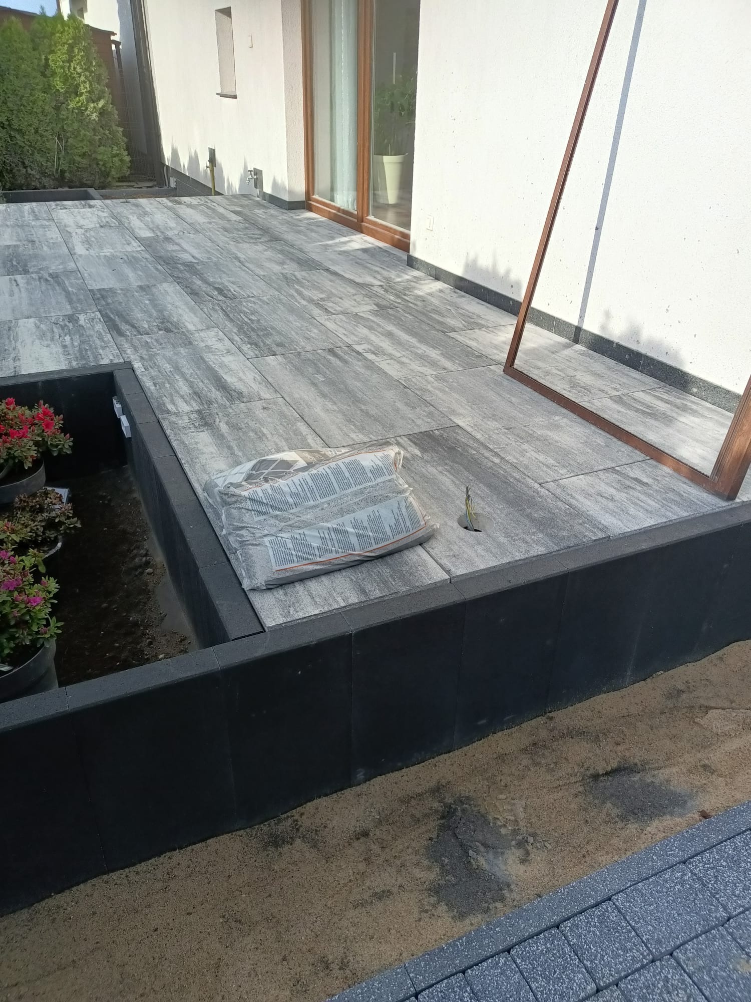 Nowoczesny taras z szarej kostki brukowej, obrzeża z czarnych betonowych elementów. W tle jasny budynek z oknami.