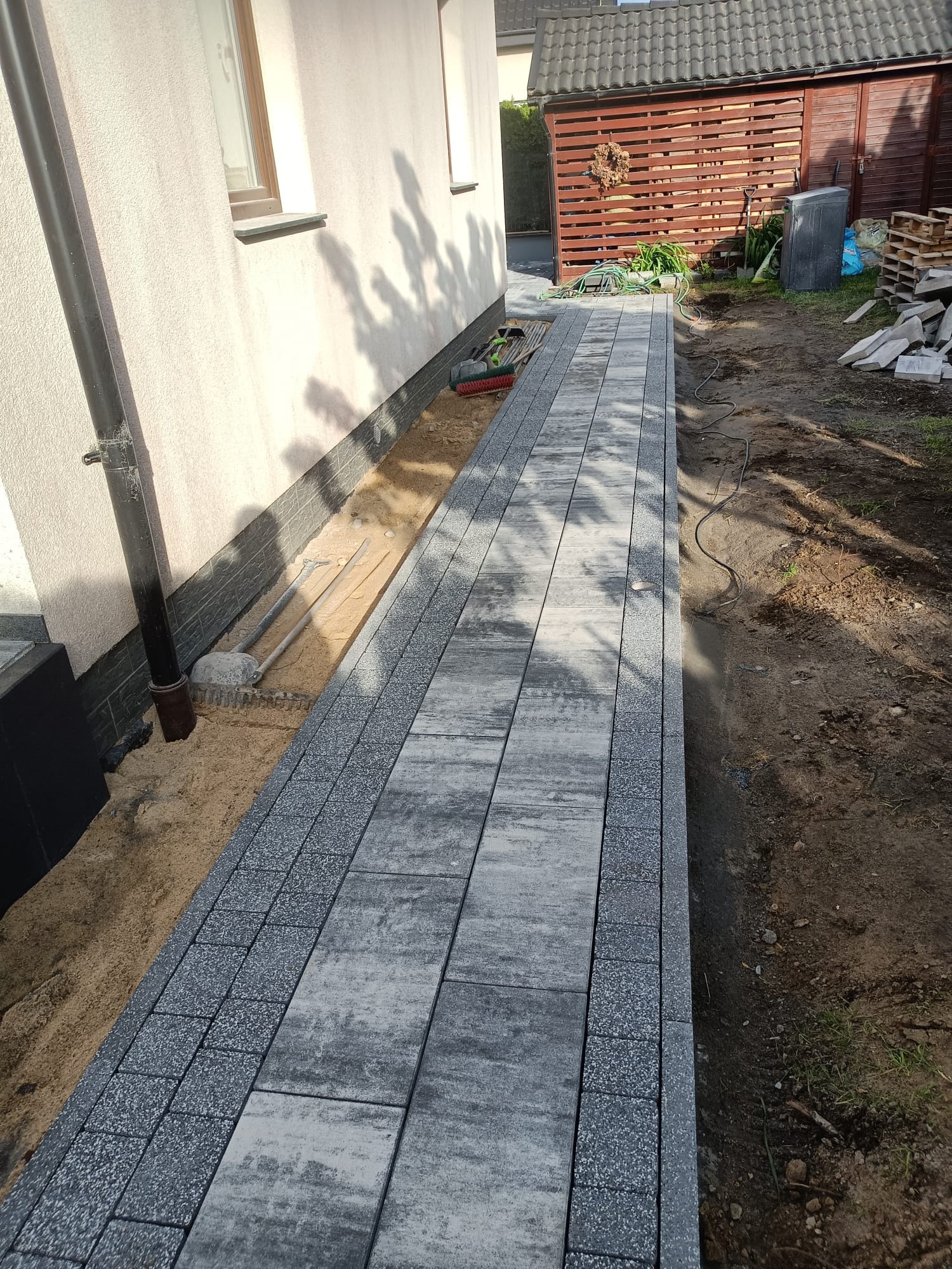 Ułożona ścieżka z szarych płyt betonowych obrzeżonych kostką, prowadząca wzdłuż budynku z widocznymi narzędziami brukarskimi i przygotowanym piaskowym podłożem.